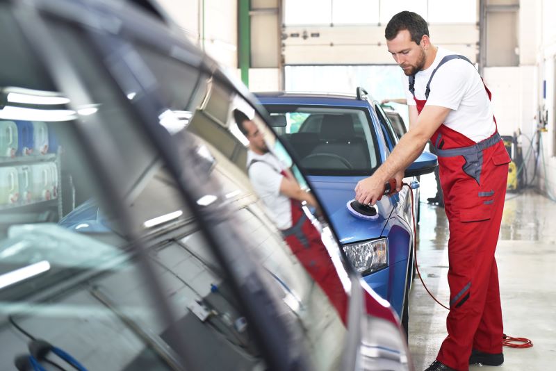 Homem vestindo macacão vermelho faz o polimento do carro com uma máquina específica em uma oficina.