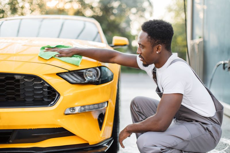 Profissional usando macacão cinza limpando um automóvel amarelo com produtos de limpeza automotiva.