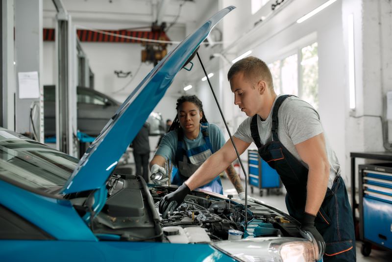 O que é manutenção preventiva: dois mecânicos usando ferramentas para consertar motor de carro com capô aberto em uma oficina.